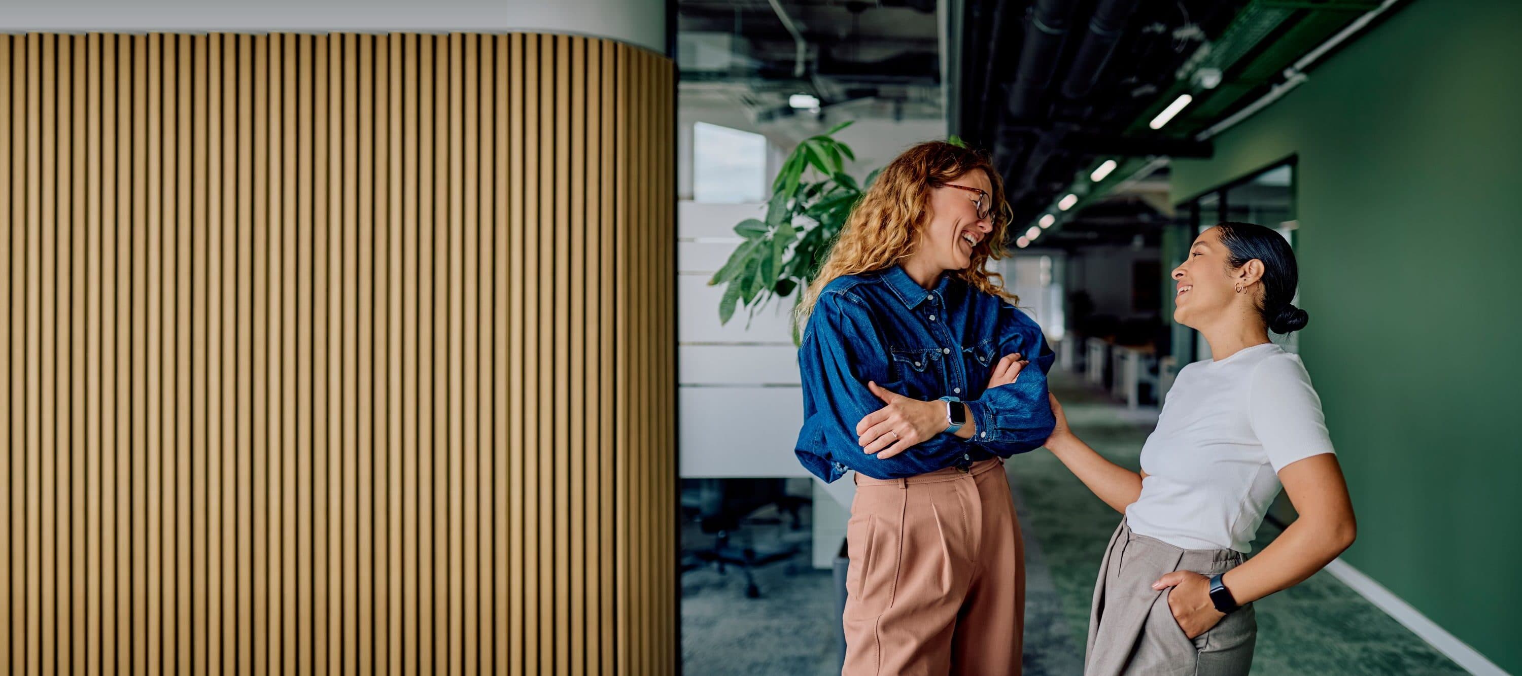 Two colleagues laugh together in an office corridor