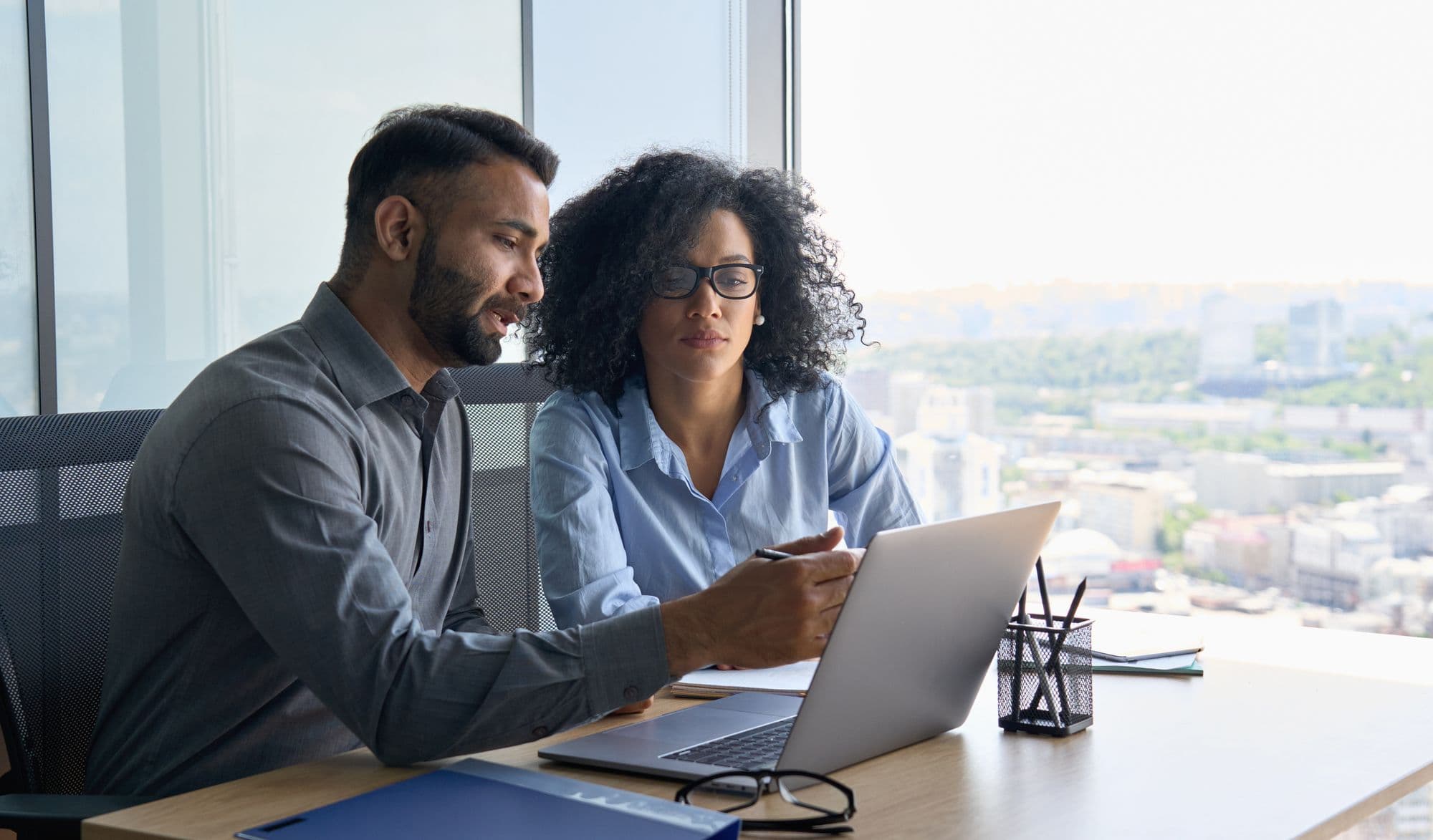 A man and a woman review details on a laptop together at a desk in an office overlooking a city