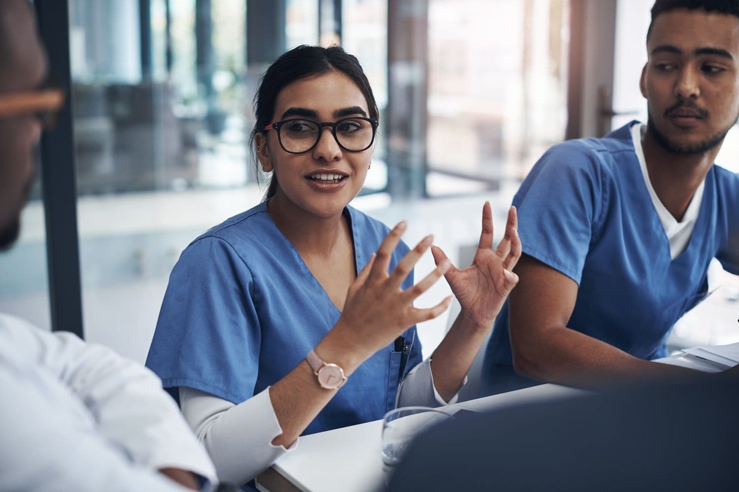 A nurse in scrubs explains something to colleagues around a hospital meeting table