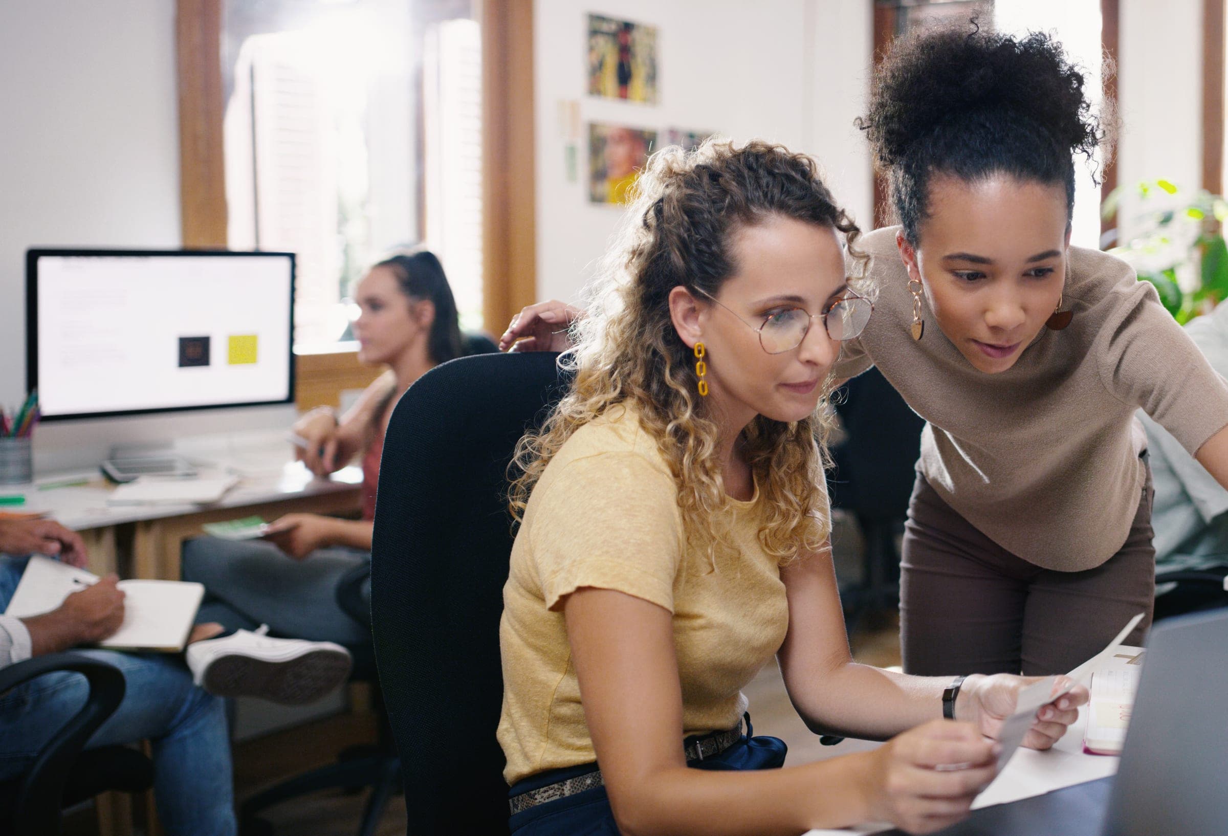 A colleague leans over a coworker’s desk to go through a printout with her in a busy open-plan office