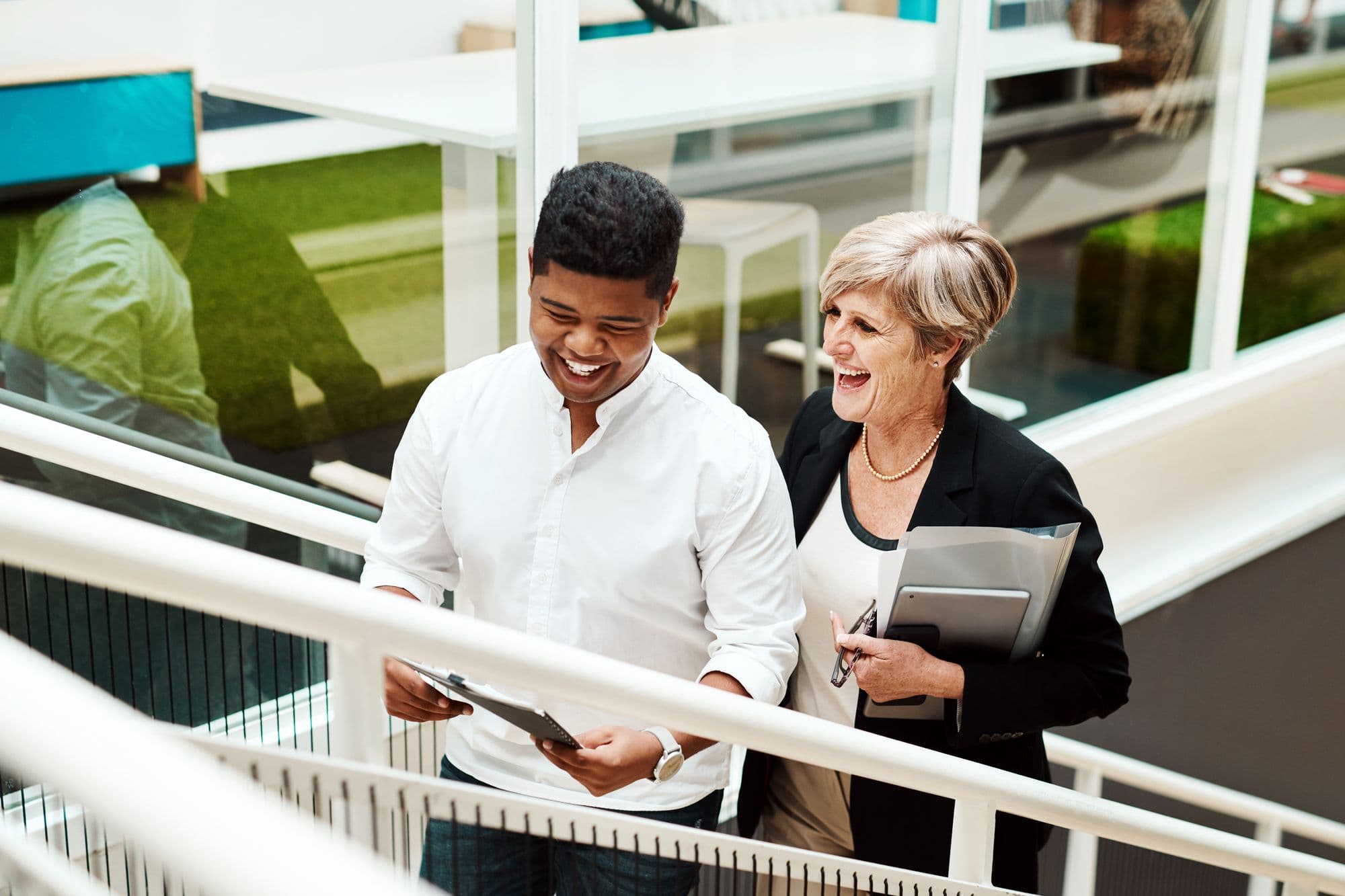 An experienced employee holding a folder and tablet laughs with a younger colleague as they walk up the office stairs