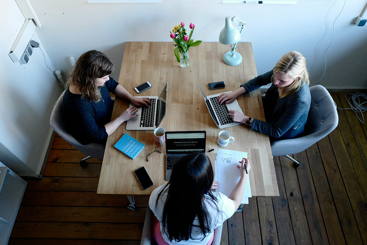 Three women sitting at a table in an office on their laptops. They're building an upskilling mentoring program.