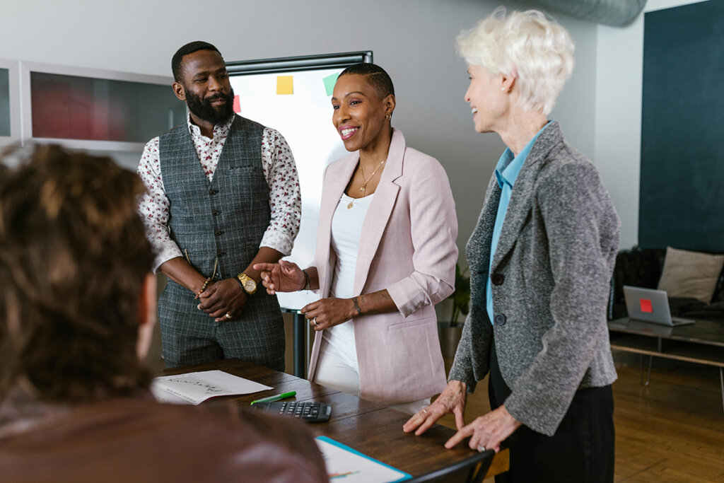 man in suit standing next to two female colleagues in an office while one of the women gives a presentation on leadership topics
