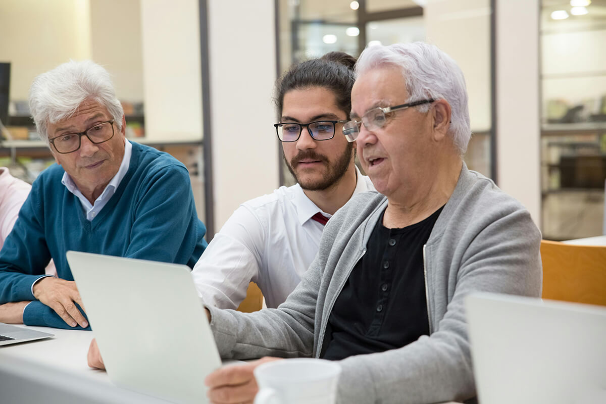 Three older men look at a laptop during a team mentoring meeting, learning about the company's new intranet system.