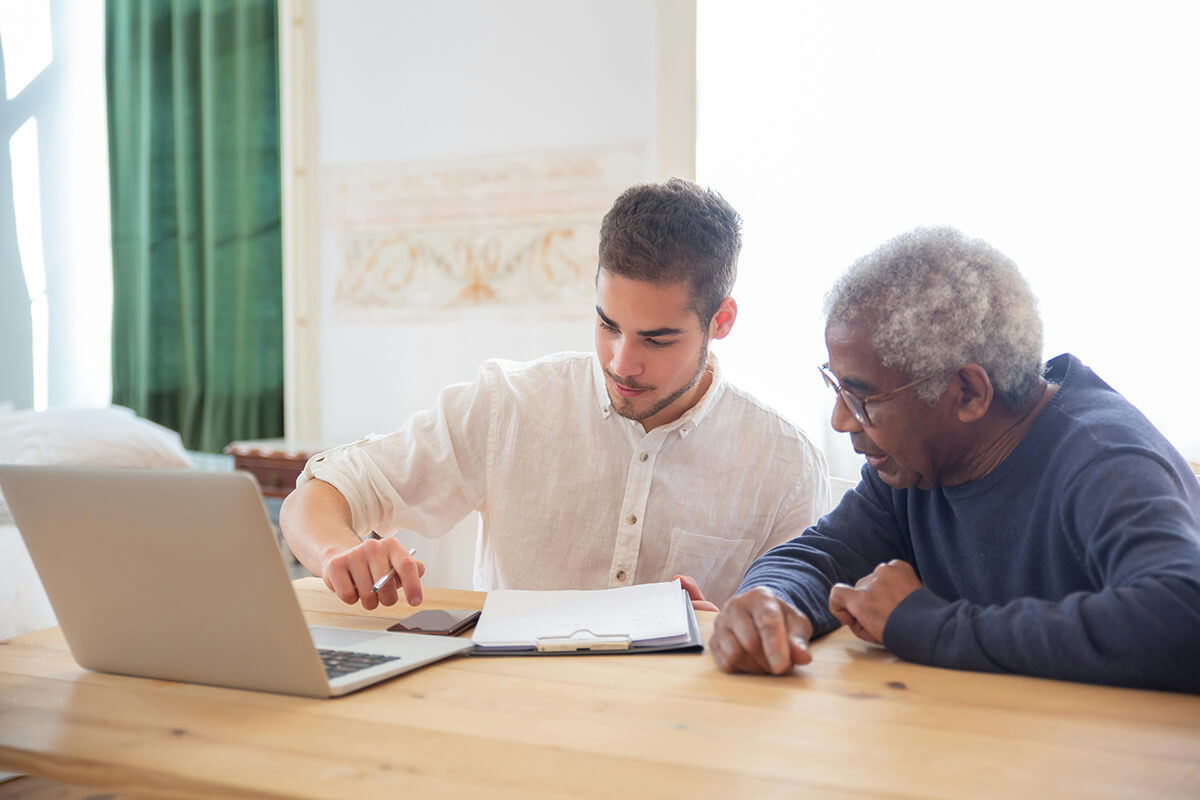 A man sitting at a desk with a male colleague showing him something on his laptop. They're talking through the 3 C's of mentoring.