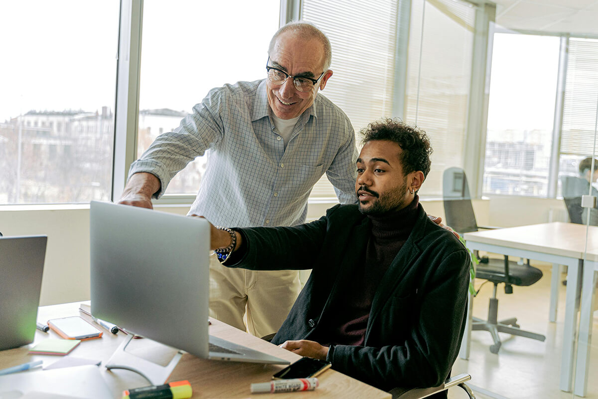 Two men in a modern office working at a desk on a laptop. Tee older man points to the screen, showing the younger colleague the 3 C's of mentorship.