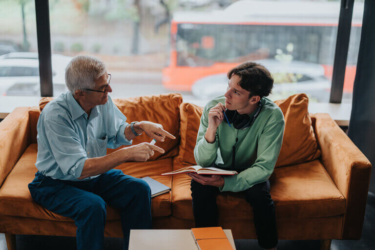 Older male colleague and younger male colleague sitting on an orange couch in a modern office during a global mentoring program discussion.