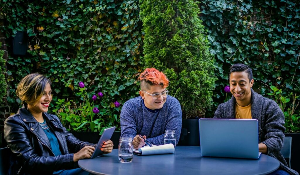 A group of three coworkers sit at an outdoor table discussing work over laptops.