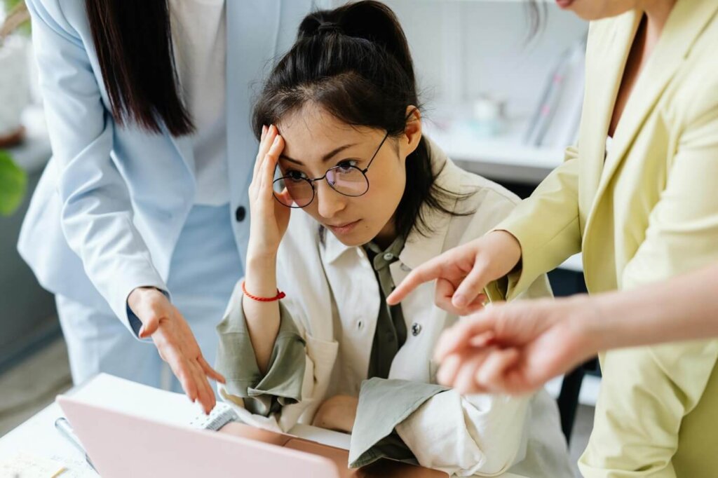 A professional looks stressed while two colleagues point at her work on a screen.